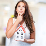 Femme pensive tenant une maquette de maison, illustrant une réflexion sur le choix d’une agence immobilière à La Rochelle.